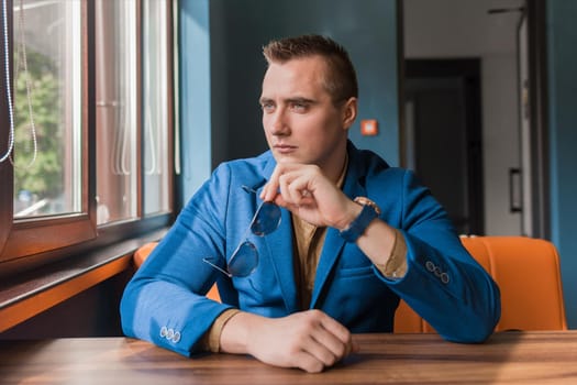 A brooding, stylish guy businessman of Caucasian attractive appearance holds glasses in his hands and looks out the window sitting at a table in a cafe.