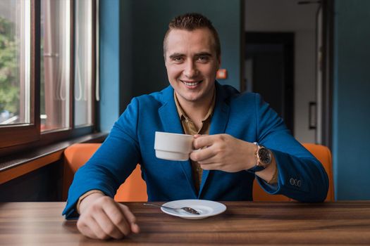 Stylish, adult attractive man of European appearance in a suit sits at a table in a cafe and drinks coffee on a lunch break.