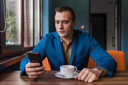 A businessman of Caucasian appearance who is keen on a mobile phone or smartphone sits at a table in a cafe on a coffee break.