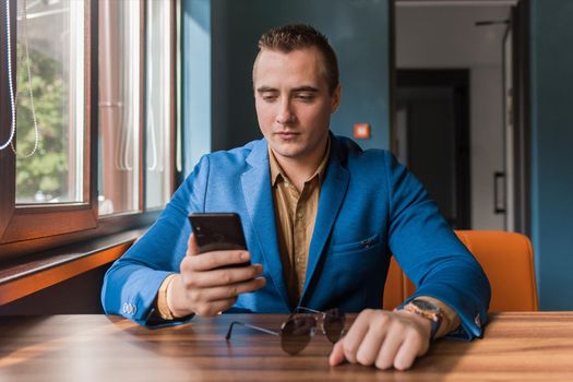 Fascinated by a mobile phone or smartphone in the hand of a stylish businessman of European appearance, in a jacket sits at a table in a cafe.