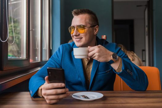 A smiling businessman of European appearance in sunglasses, a jacket and shirt sits at a table in a cafe on a coffee break, holds a smartphone or mobile phone in his hand and looks out the window.