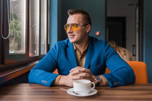 A positive smiling businessman a stylish portrait of Caucasian appearance in sunglasses, sits idly by at a table on a coffee break in a cafe background and looks out the window.