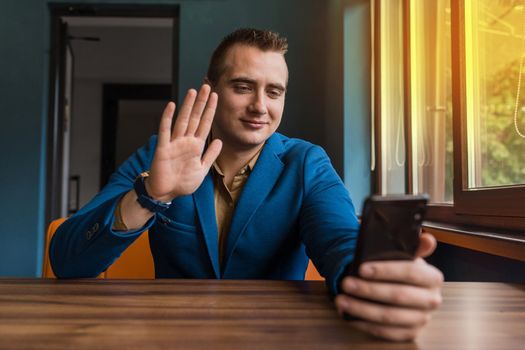 A young stylish guy, businessman of European appearance portrait sits at a table in a suit and talks via video communication in a smartphone transmitting greetings with a hand gesture.