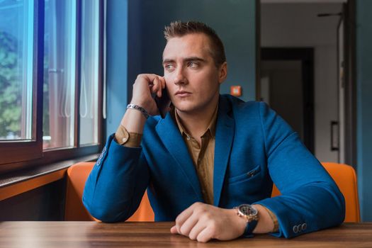 An adult stylish, businessman of European appearance portrait sits in a blue jacket at a table in a cafe and talks on a smartphone looking out the window.