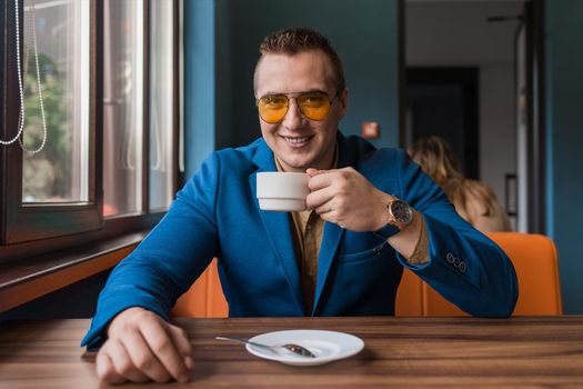 A positive smiling businessman a stylish portrait of Caucasian appearance in sunglasses, jacket and shirt, sits at a table on a coffee break in a cafe background.