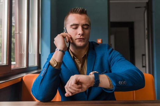 A man of stylish European appearance portrait of a businessman in a jacket talks on a mobile phone or smartphone, and looks at the time on the clock on his hand in a cafe.