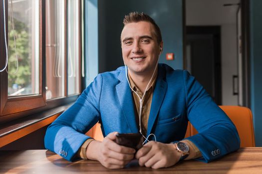 Businessman, a stylish, handsome smiling guy of Caucasian appearance portrait in a blue jacket spends time in a smartphone or mobile phone sitting at a table in the cafe.