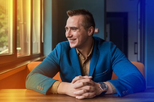 A smiling positive, young man of European appearance portrait businessman in jacket and shirt sits idly by at a table in a cafe and looks out the window.