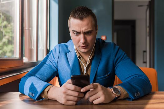 Businessman, a stylish, surprised handsome guy of Caucasian appearance portrait in a blue jacket spends time in a smartphone or mobile phone sitting at a table in the cafe.
