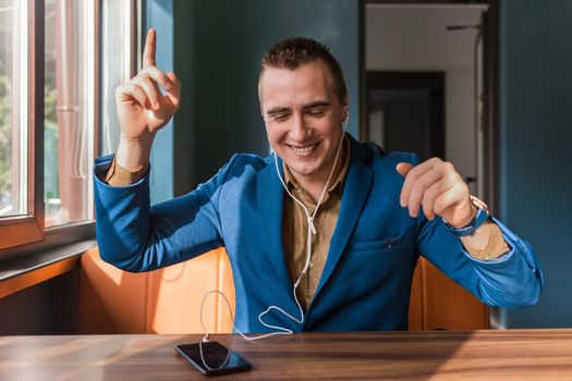 A handsome, happy, dancing businessman of European appearance stylish portrait, holds a phone in his hand and listens to music in headphones sitting at a table in a cafe.