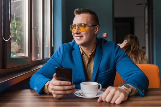 A smiling guy businessman of European appearance in sunglasses, a jacket and shirt sits at a table in a cafe on a coffee break, holds a smartphone or mobile phone in his hand and looks out the window.