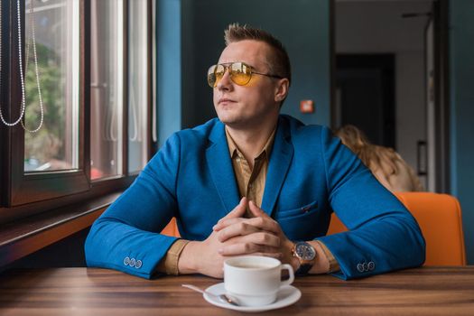 A brooding businessman a stylish portrait of Caucasian appearance in sunglasses, sits idly by at a table on a coffee break in a cafe and looks out the window.