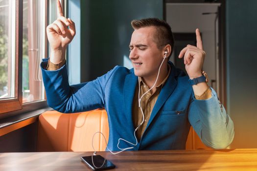 A handsome, funny dancing businessman of European appearance stylish portrait, holds a phone in his hand and listens to music in headphones sitting at a table in a cafe.