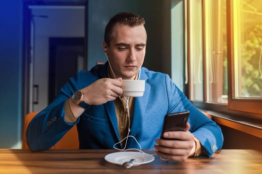 A young handsome guy businessman in blue jacket, of European appearance portrait, uses a smartphone or mobile phone sitting in a cafe at a table on a coffee break.