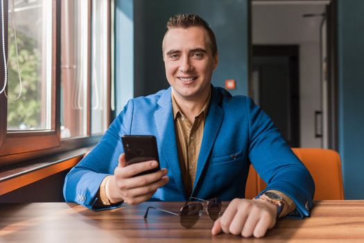 Businessman, a stylish, handsome guy of Caucasian appearance portrait in a suit and shirt spends time in a smartphone or mobile phone sitting at a table in the cafe.