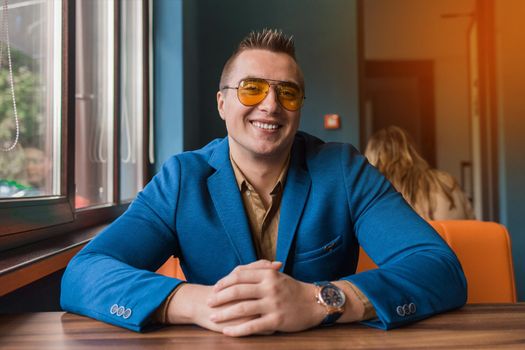 Cute positive smiling businessman of Caucasian appearance stylish portrait in sunglasses, jacket and shirt, sitting at a table in a cafe idly by.