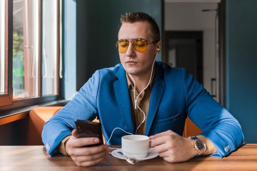 A businessman a stylish portrait of Caucasian appearance in sunglasses, blue jacket, sits at a table on a coffee break in mobile phone or cellphone a cafe background.