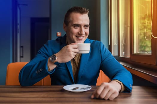 A stylish, positive businessman of Caucasian appearance, a man in a jacket and shirt sits at a table in a cafe on a coffee break and looks away or out the window.