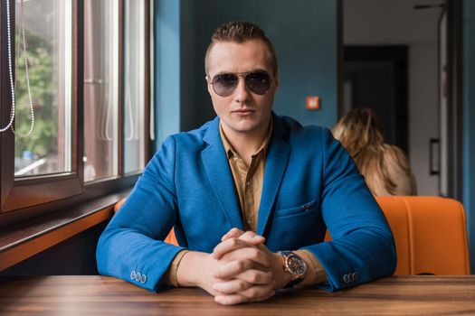 A young serious businessman of European appearance stylish portrait with glasses sits at a table in a cafe idly by.