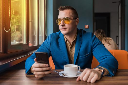 A sad guy businessman of European appearance in sunglasses, a blue jacket sits at a table in a cafe on a coffee break, holds a smartphone in his hand and looks out the window.