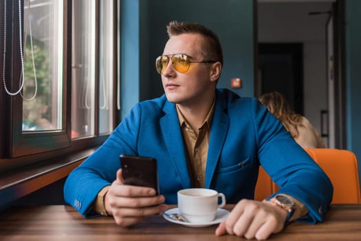 A guy businessman of European appearance in sunglasses, a jacket and shirt sits at a table in a cafe on a coffee break, holds a smartphone or mobile phone in his hand and looks out the window.
