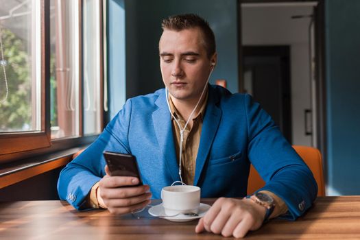 A stylish guy businessman of European appearance in a jacket and shirt sits at a table in a cafe on a coffee break, holds a smartphone or mobile phone in his hand.