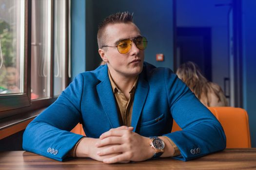 A brooding businessman of European appearance stylish portrait in sunglasses, a blue suit sits at a table in a cafe idly by and looks away.