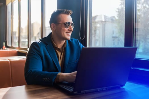 A business man happy smiling businessman a stylish portrait of Caucasian appearance in a jacket and shirt, sunglasses works in a laptop or computer, sitting at a table by the window in a cafe.