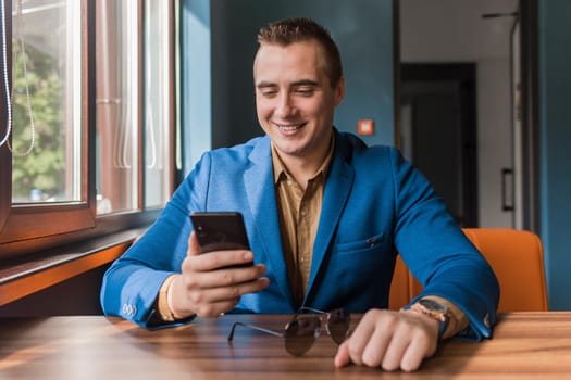 Smiling happy by a mobile phone or smartphone in the hand of a stylish businessman of European appearance, in a jacket sits at a table in a cafe.