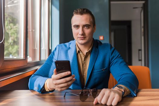 Businessman, a stylish, handsome guy of Caucasian appearance portrait in a blue suit and brown shirt spends time in a smartphone or mobile phone sitting at a table in an institution.