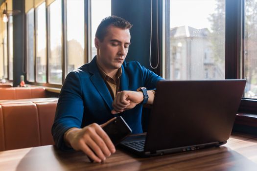 A business serious man businessman a stylish portrait of Caucasian appearance in a jacket works in a laptop or computer, sitting at a table by the window in a cafe and holds mobile phone in hand.