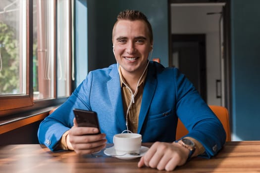 A stylish guy happy smiling businessman of European appearance in a jacket and shirt sits at a table in a cafe on a coffee break, holds a smartphone or mobile phone in his hand.