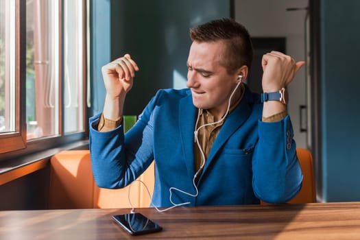 A handsome, happy, funny businessman of European appearance stylish portrait, holds a phone in his hand and listens to music in headphones sitting at a table in a cafe.