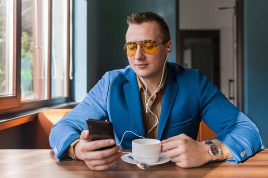 A businessman a stylish portrait of Caucasian appearance in sunglasses, suit style, sits at a table on a coffee break in smartphone and headphones, listen to music on cellphone or mobile in a cafe background.