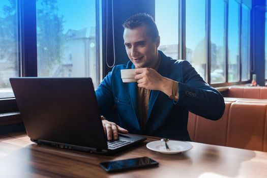 A business smiling man businessman a stylish portrait of Caucasian appearance in a jacket, works in a laptop or computer, drink coffee on coffee break, sitting at a table by the window in a cafe.