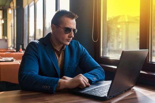 A business man businessman a stylish portrait of Caucasian appearance in a blue jacket and sunglasses works in a laptop or computer, sitting at a table by the window in a cafe.