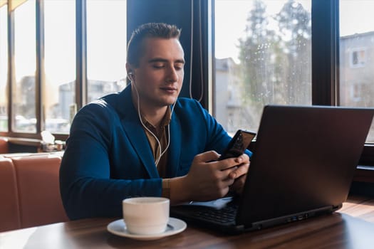 A business man stylish businessman in an attractive European-looking suit works in a laptop, listens to music with headphones and drinks coffee sitting at a table in a cafe by the window.