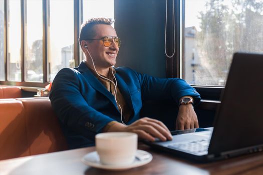 A business positive man smiling businessman a stylish of Caucasian appearance, in sunglasses, works in a laptop or computer, sitting at a table by the window in a cafe and listening to music.