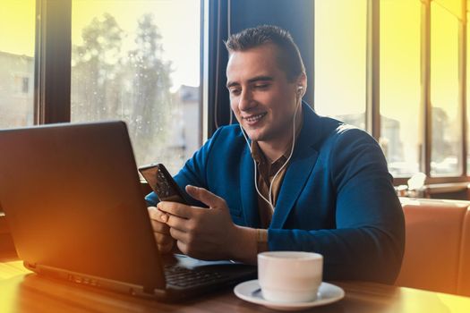 A business man stylish positive businessman in an attractive European-looking suit works in a laptop, listens to music with headphones and drinks coffee sitting at a table in a cafe by the window.