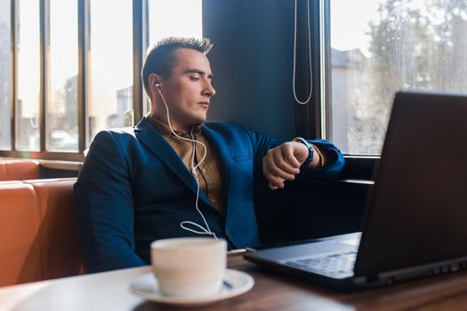 A business serious man businessman a stylish of Caucasian appearance in a jacket, works in a laptop or computer, sitting at a table by the window in a cafe and looks at the time on a wrist watch.