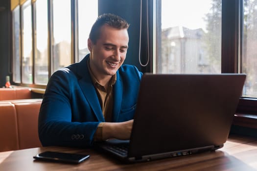 A business smiling man happy businessman a stylish portrait of Caucasian appearance in a jacket works in a laptop or computer, sitting at a table by the window in a cafe.