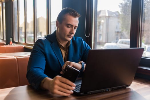 A business serious man businessman a stylish portrait of Caucasian appearance in a jacket works in a laptop or computer, sitting at a table by the window in a cafe and holds mobile phone in hand.