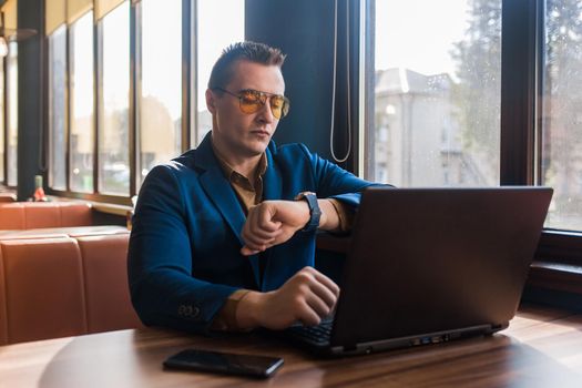 A business serious man businessman a stylish of Caucasian appearance in a jacket and sunglasses, works in a laptop or computer, sitting at a table by the window in a cafe and holds mobile phone.