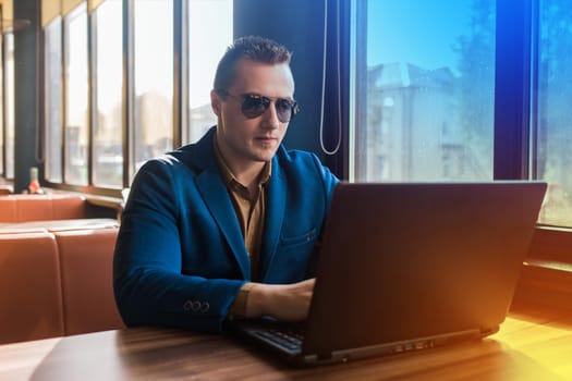 A business man businessman a stylish portrait of Caucasian appearance in a blue jacket and sunglasses works in a laptop or computer, sitting at a table by the window in a cafe.