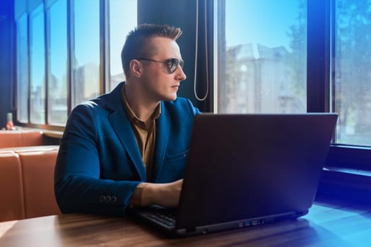 A business man businessman a stylish portrait of Caucasian appearance in a jacket and sunglasses works in a laptop or computer, sitting at a table by the window in a cafe.