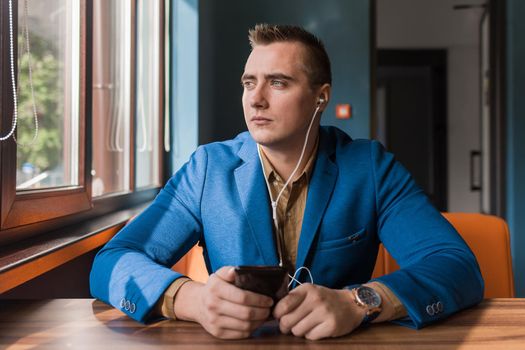 Businessman, a stylish, handsome guy of Caucasian appearance portrait in a suit spends time in a smartphone or mobile phone sitting at a table in the cafe and looking out the window background.