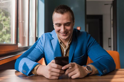 Businessman, a stylish, cute handsome guy of Caucasian appearance portrait in a blue jacket spends time in a smartphone or mobile phone sitting at a table in the cafe.
