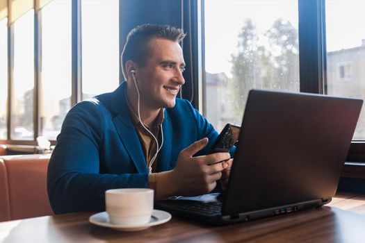 A business man stylish positive smiling businessman in an attractive European-looking suit works in a laptop, listens to music with headphones and sitting at a table in a cafe by the window.