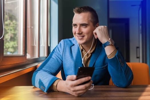 A handsome smiling businessman of European appearance stylish portrait, holds a phone in his hand and listens to music in headphones sitting at a table in a cafe.