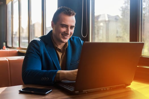 A business smiling man happy businessman a stylish portrait of Caucasian appearance in a jacket works in a laptop or computer, sitting at a table by the window in a cafe.